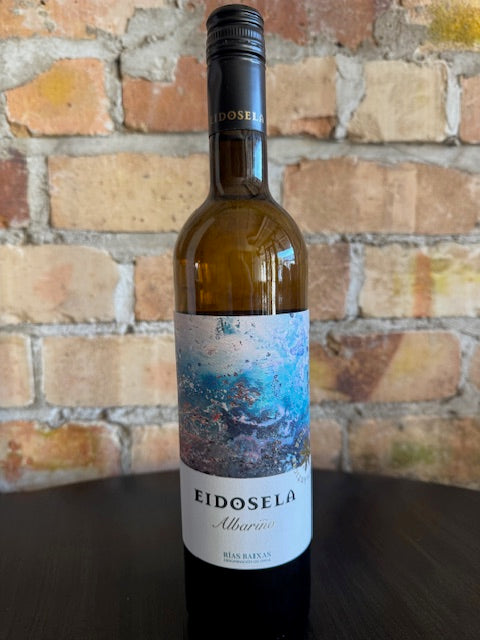 Wine bottle with label against a brick wall background.  The label has text "Eidosela Albarino" below an abstract pretty blue watercolour picture.