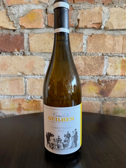 Wine bottle with a label on a brick surface.  The label shows an old black and white picture of people enjoying wine and the big text reads Famillle Guilhem Chardonnay