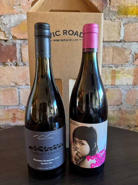 Two wine bottles with labels on a cardboard box against a brick wall background