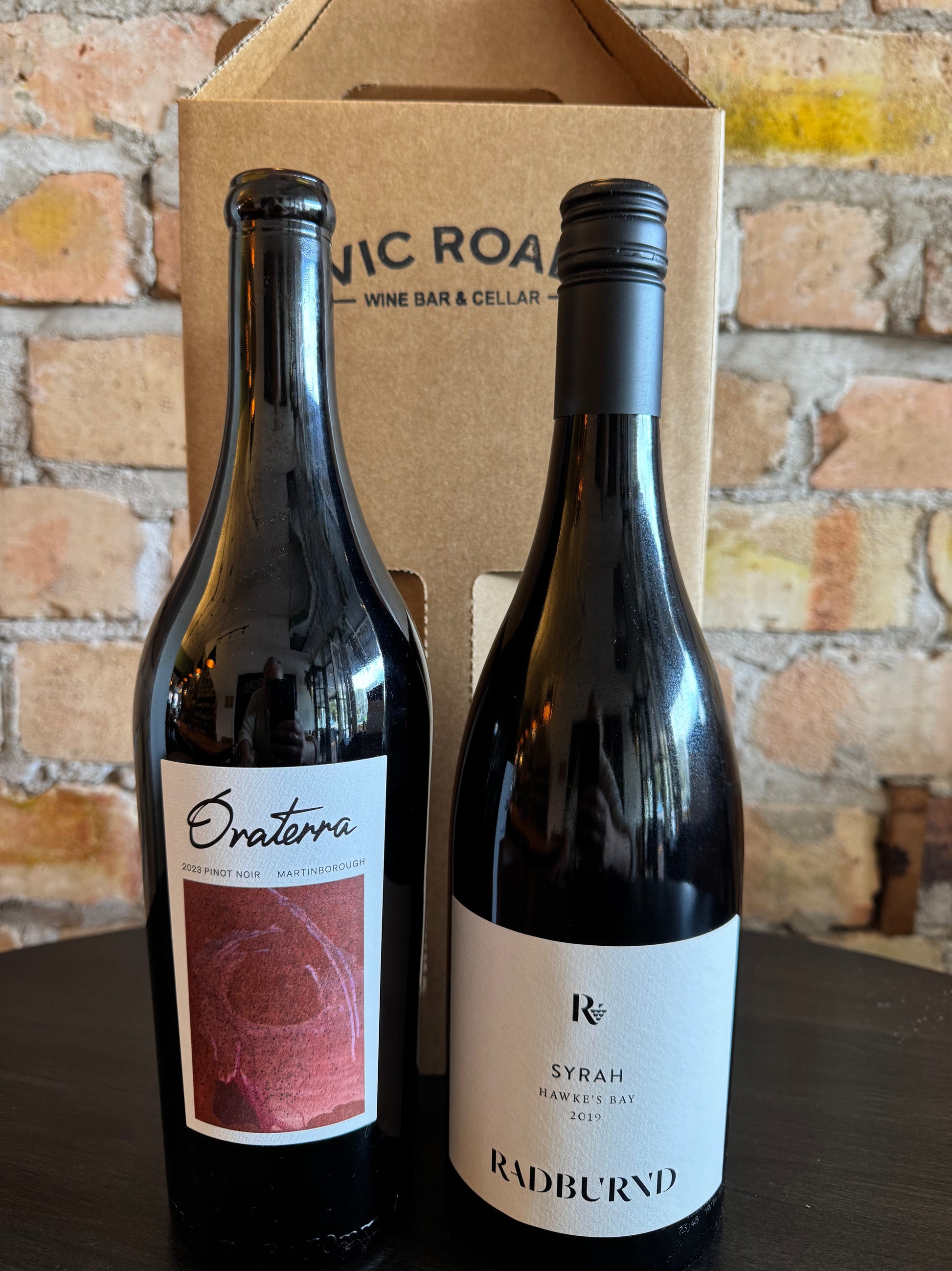 Two wine bottles, one labeled 'Oraterra' and the other 'Radburnd', in front of a cardboard box with 'Vic Road Wine Bar & Cellar' branding on a brick wall background.