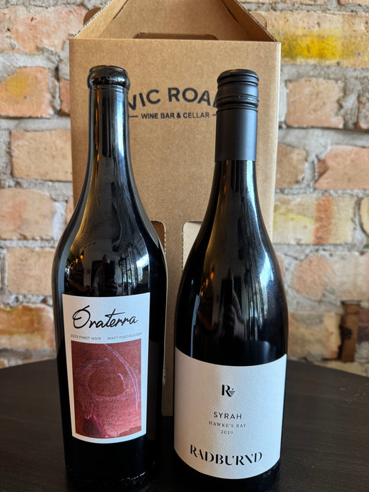 Two wine bottles, one labeled 'Oraterra' and the other 'Radburnd', in front of a cardboard box with 'Vic Road Wine Bar & Cellar' branding on a brick wall background.