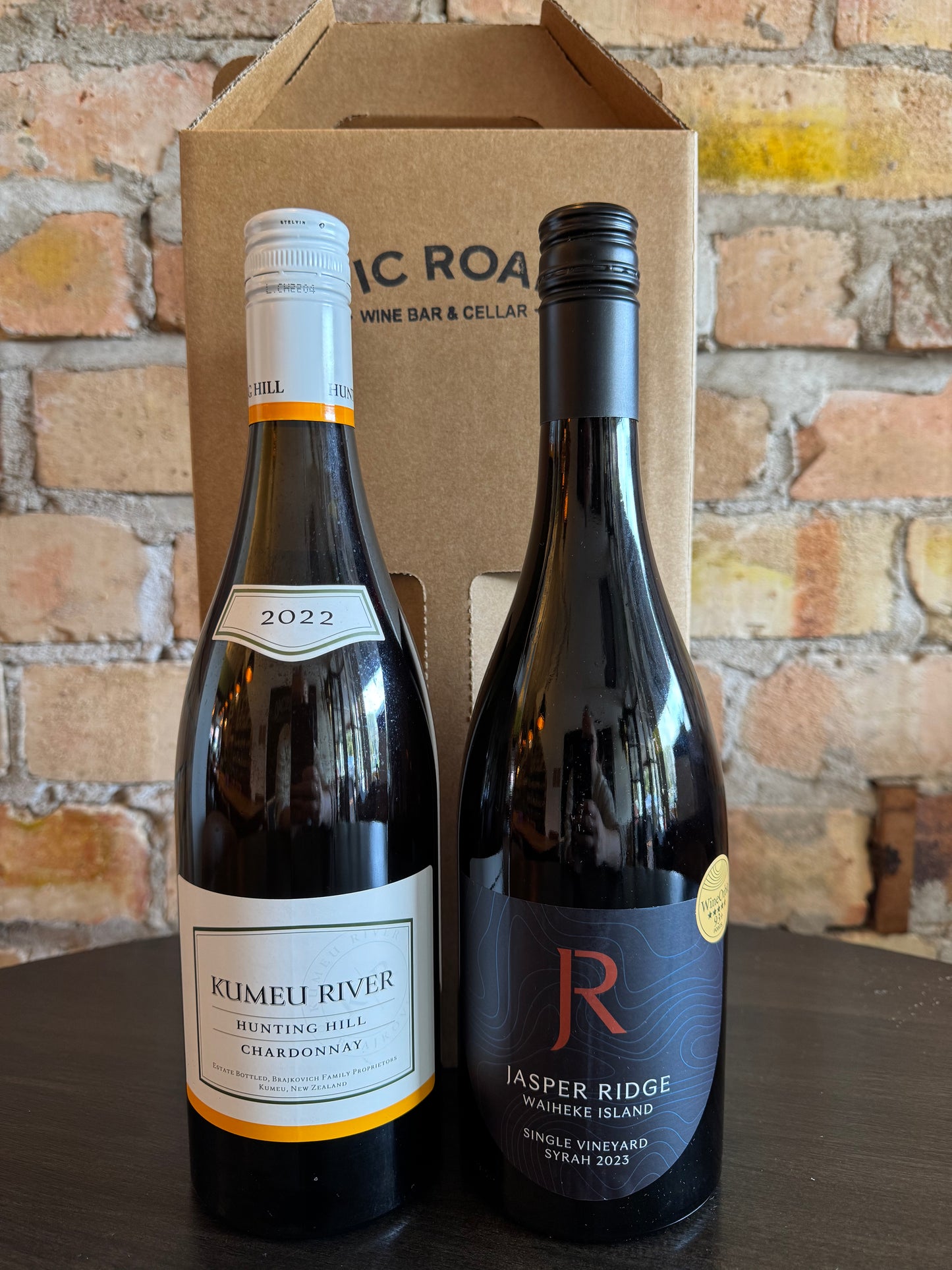Two wine bottles, one labeled 'Kumeu River' and the other 'Jasper Ridge', in front of a cardboard box with 'Vic Road Wine Bar & Cellar' branding.